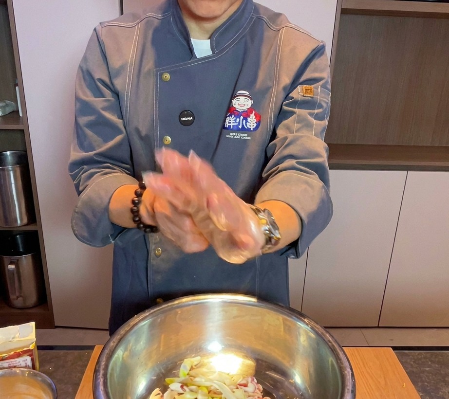 Chef wearing a clear glove mixing raw pork belly and aromatics in a large metal bowl.