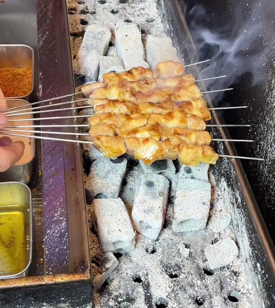 Pork belly skewers turning golden-brown as they sizzle over a hot, smoky charcoal grill.