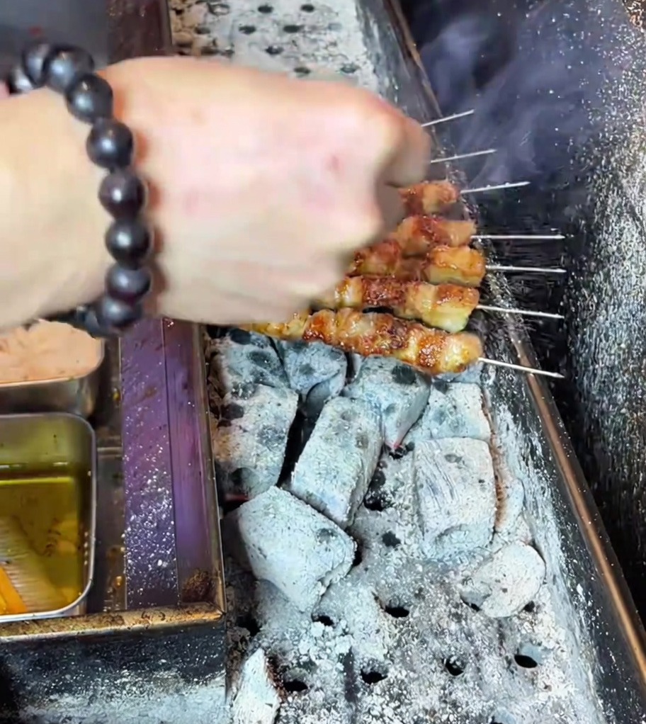 Hand sprinkling seasoning powder over perfectly grilled, golden-brown pork belly skewers on the charcoal grill.