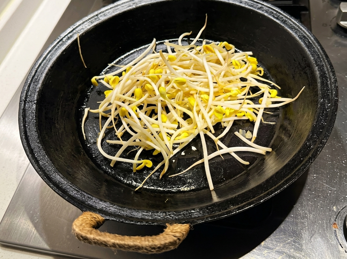 Cleaned, fresh soybean sprouts being spread out in a pre-heated, oiled black skillet.
