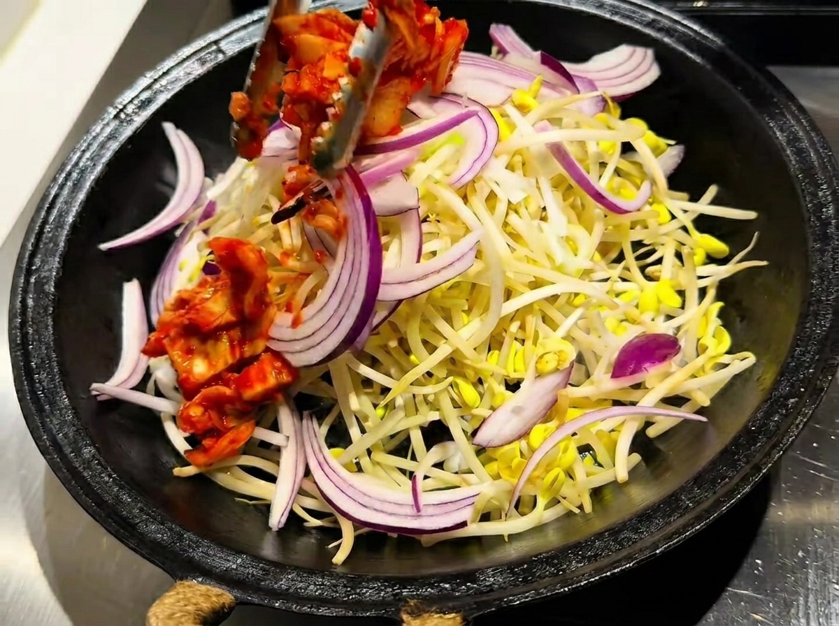 A pan containing a layer of bean sprouts topped with vibrant red kimchi and sliced raw onions.