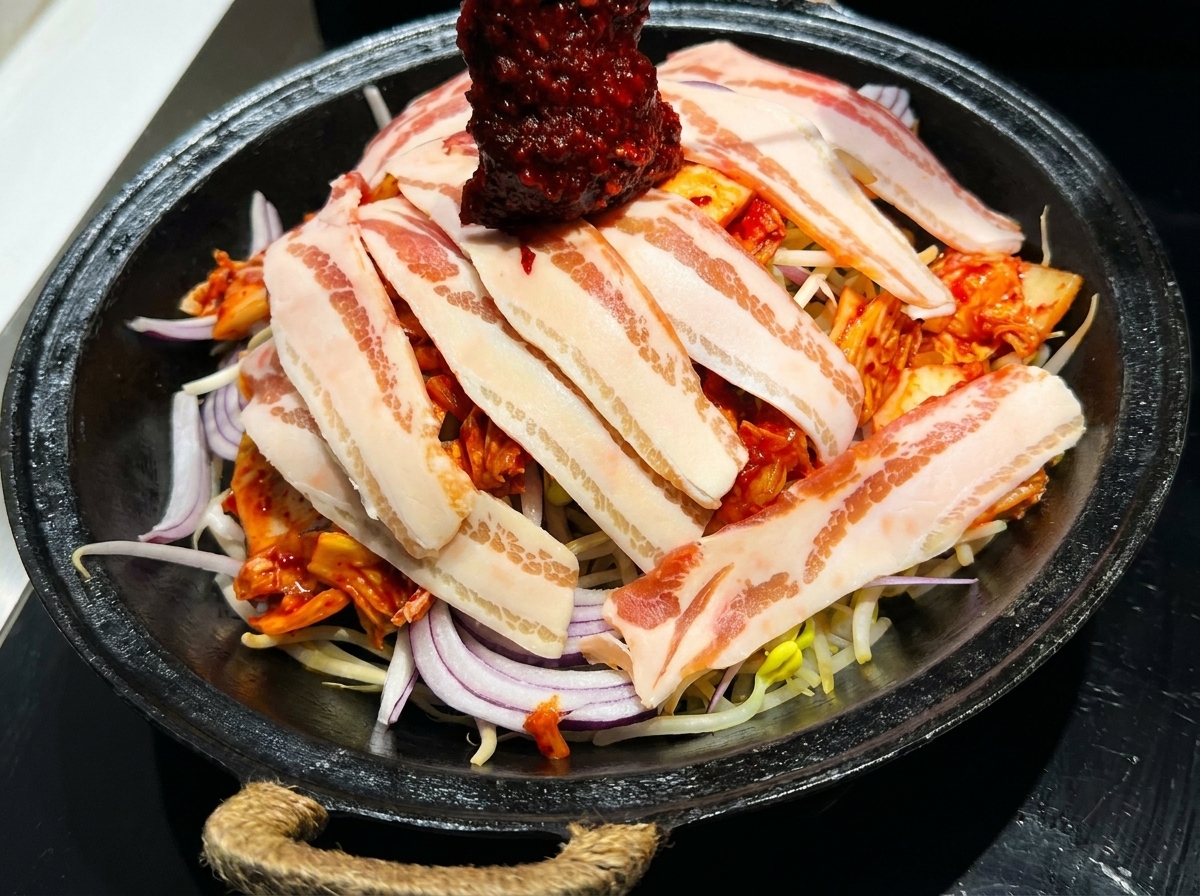 A vibrant red Korean stir-fry sauce being spread over a pan of layered pork belly and fresh vegetables.
