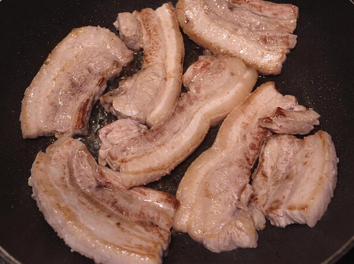 Several slices of pork belly searing in a pan, with surfaces deeply browned.
