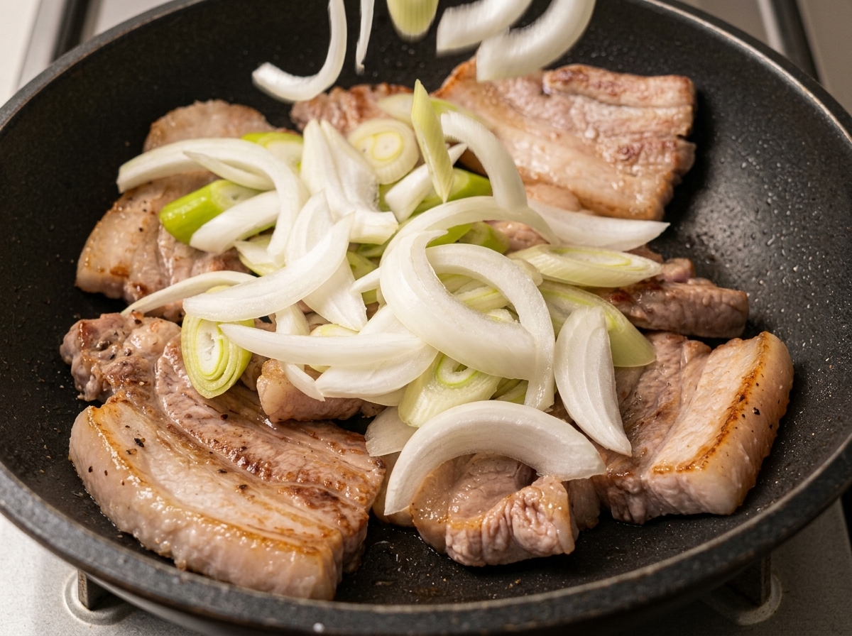 Sliced white onions and green onions scattered over browned pork belly slices in a pan.