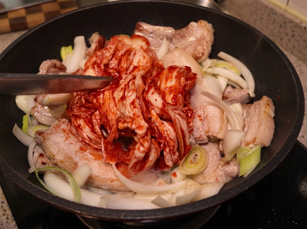 A large, whole piece of sour kimchi being placed with tongs onto the seared pork belly and onions in a pan.