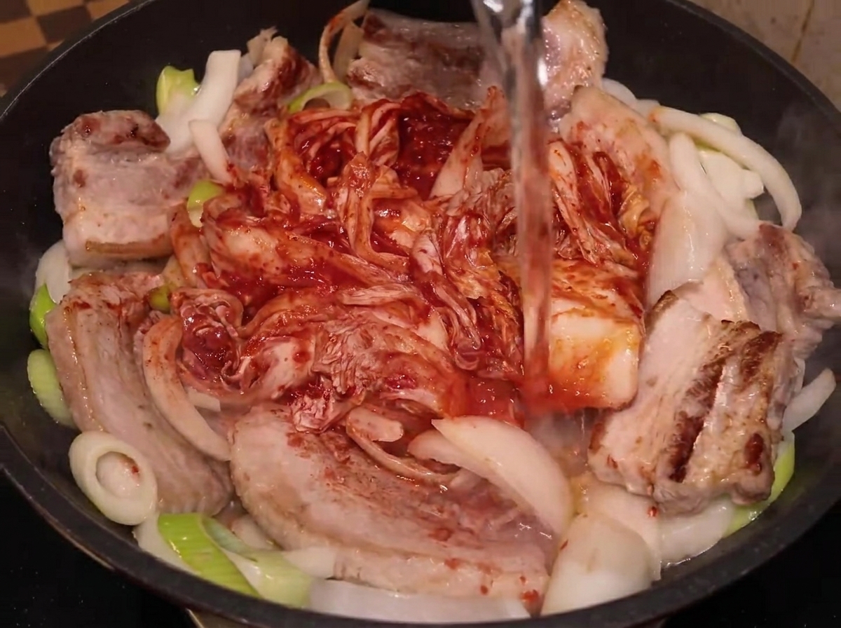 Water being poured from a plastic bottle into a pan containing pork belly, onions, and whole kimchi.