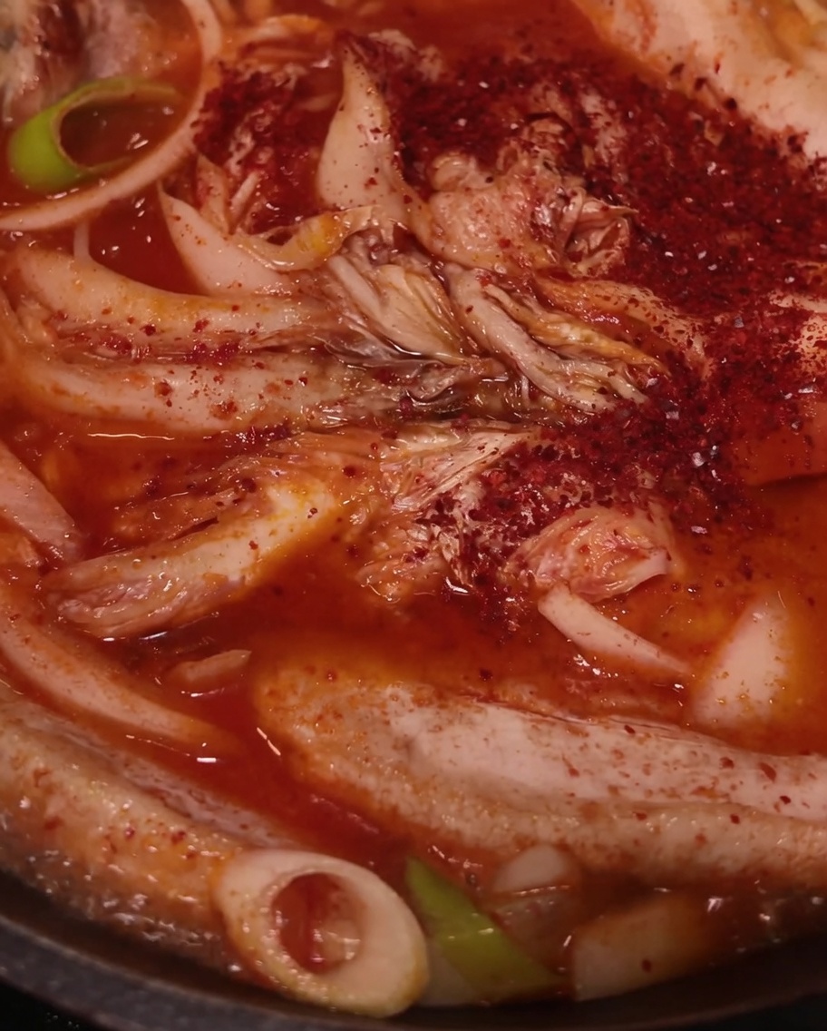 Red chili powder being sprinkled over the pork, kimchi, and onions in the pan.