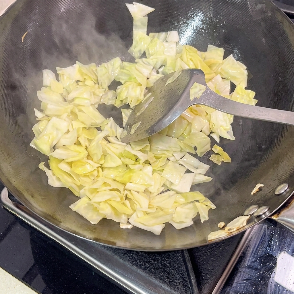 A metal spatula tossing pale green pickled cabbage in a hot black wok, with visible steam rising from the pan.