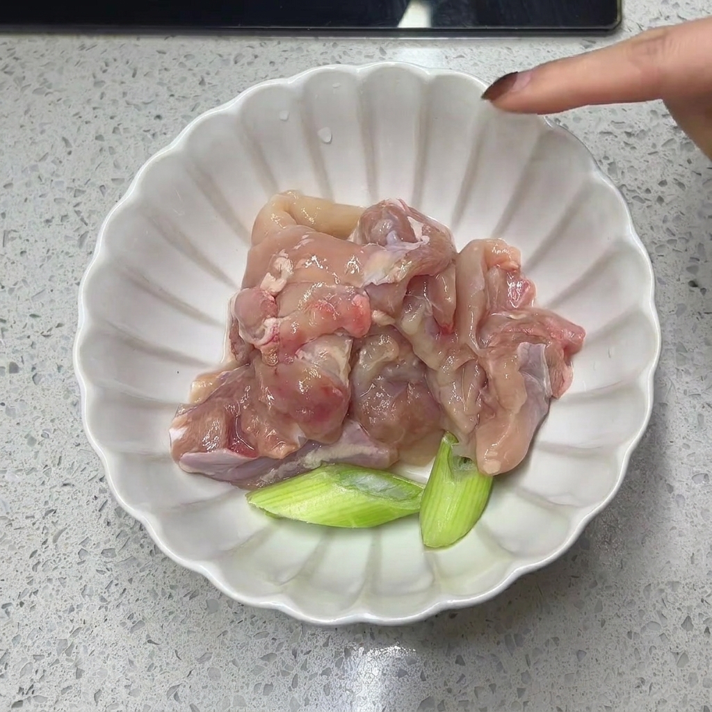 Raw, deboned chicken thigh meat and green onion segments resting in a white scalloped bowl.