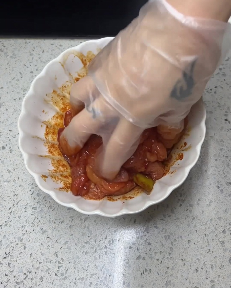 A gloved hand massaging a red, spiced marinade into raw chicken pieces in a white bowl.