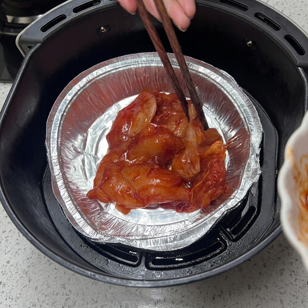 Wooden chopsticks arranging marinated raw chicken pieces inside an aluminum foil tray placed in an air fryer basket.