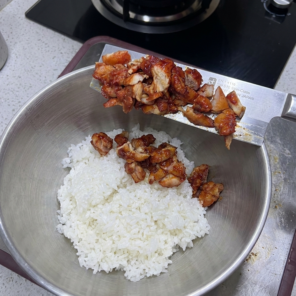 Chopped cooked chicken being slid off a wide knife into a large metal bowl filled with fluffy white rice.