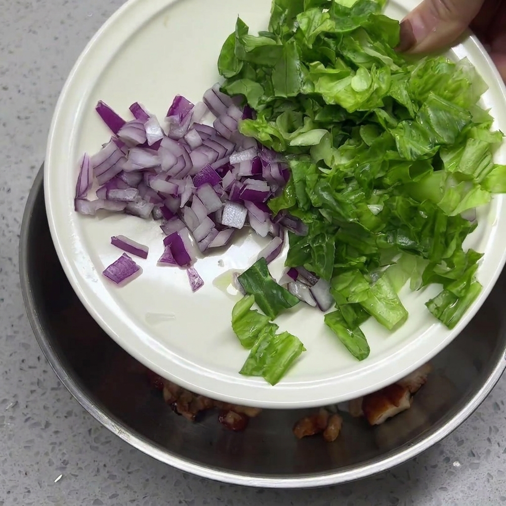 Minced red onion and shredded green lettuce being poured from a white plate into a bowl containing chicken and rice.