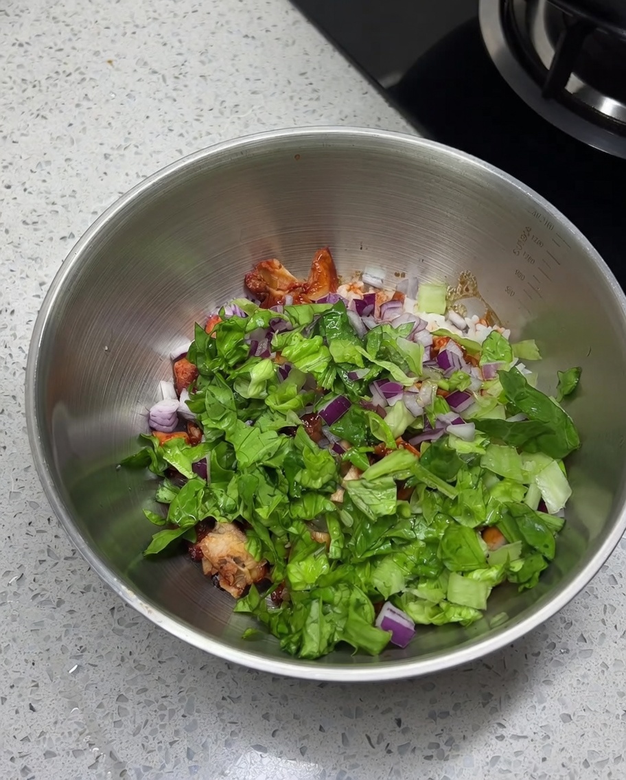 Close-up of salt, cumin powder, and chili powder being sprinkled onto the mixture of chicken, rice, and fresh vegetables in a stainless steel bowl.