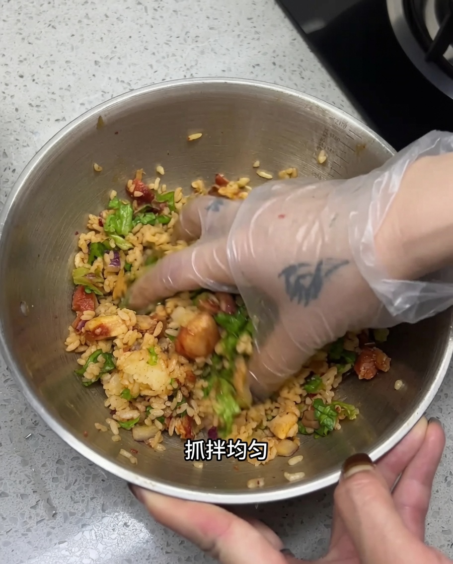 A gloved hand mixing the rice, chopped chicken, and vegetables in a large stainless steel bowl to ensure all components and seasonings are well combined.