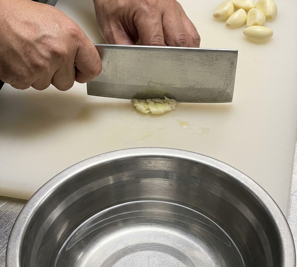 A close-up of a chefs knife chopping garlic and ginger on a white cutting board.