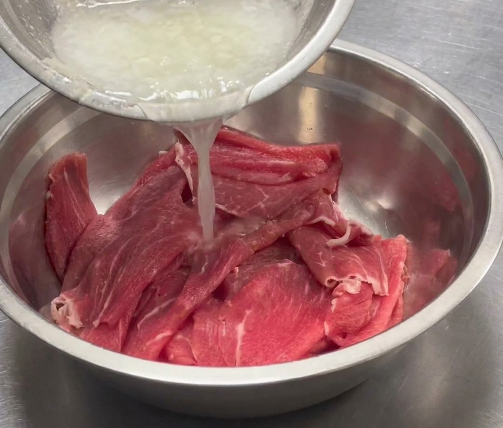 Aromatic puree being poured into a large steel bowl containing sliced beef.