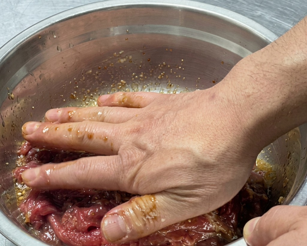 Hands thoroughly mixing the raw beef with the marinade inside the bowl.