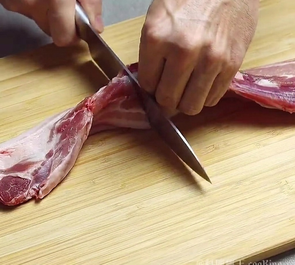 A person using a chefs knife to cut a piece of raw pork belly into thick strips on a wooden board.