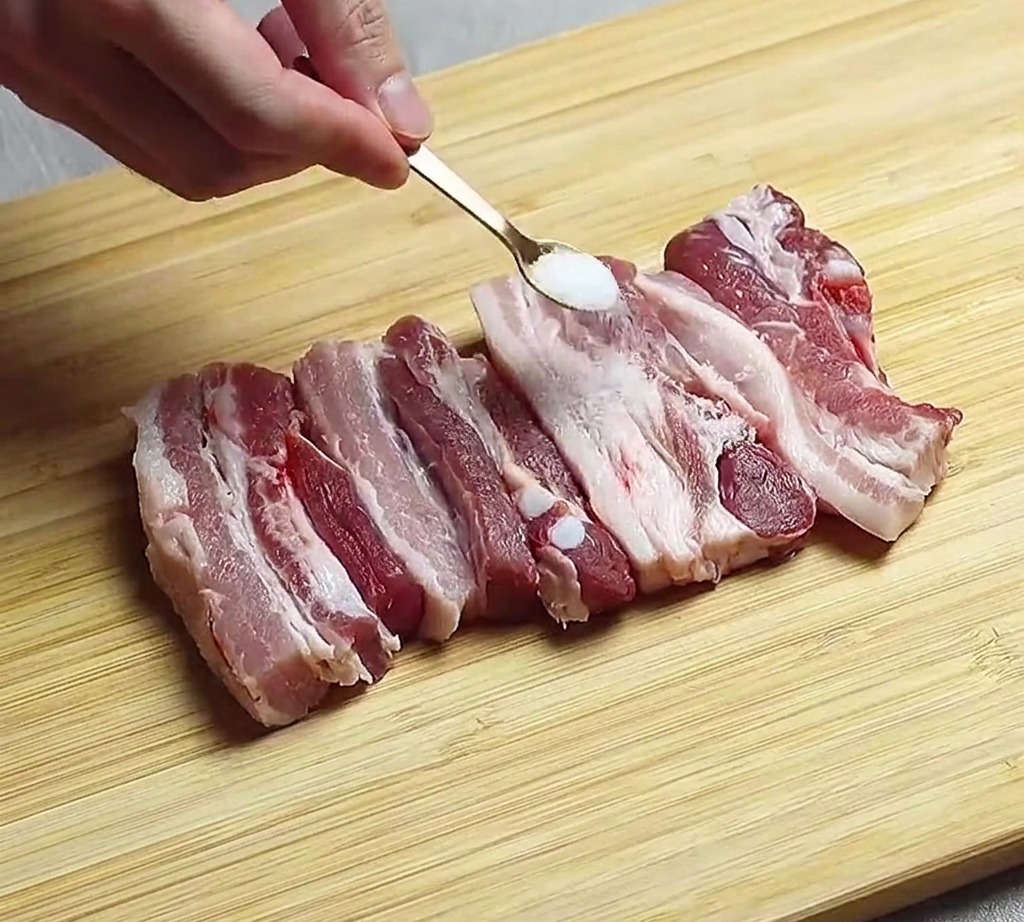 Slices of raw pork belly laid out on a wooden board being seasoned with a sprinkle of salt.