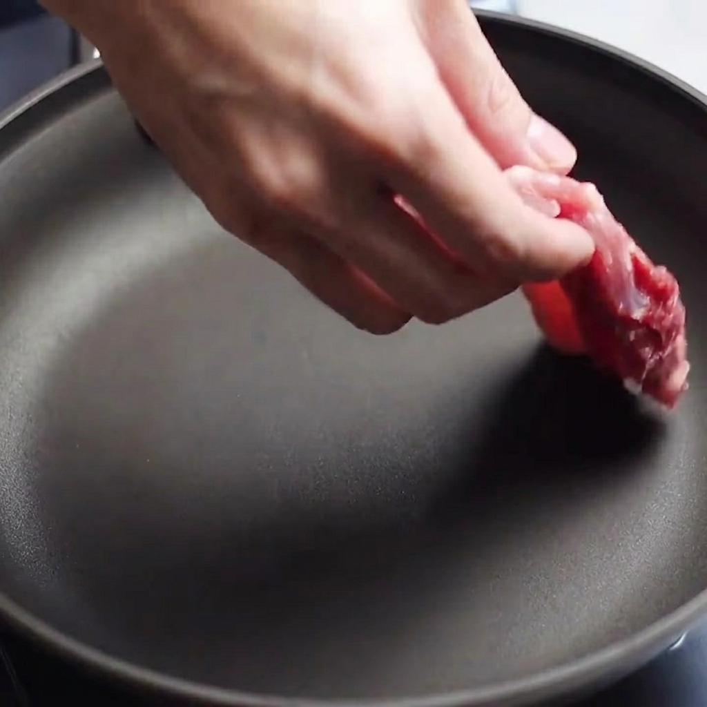 Raw, seasoned pork belly strips being placed into a preheated frying pan.