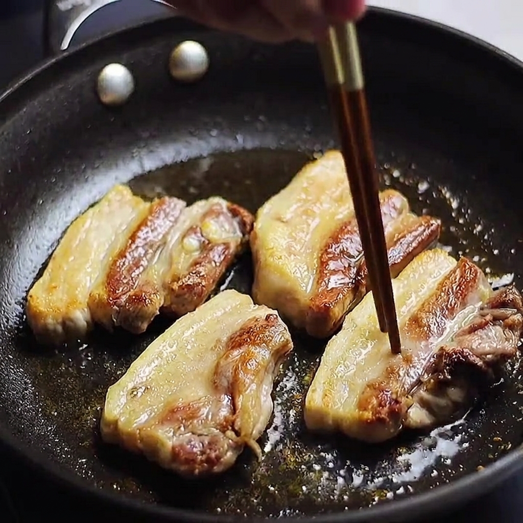 Golden-brown pork belly strips frying in a pan, with a chopstick being used to check if the meat is cooked.