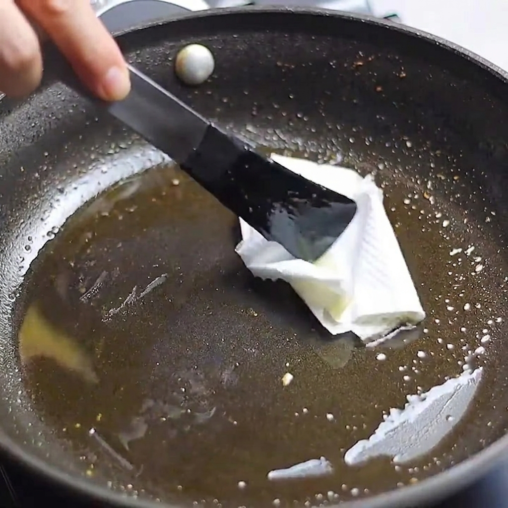 A kitchen paper towel held by tongs being used to wipe excess oil from a frying pan.