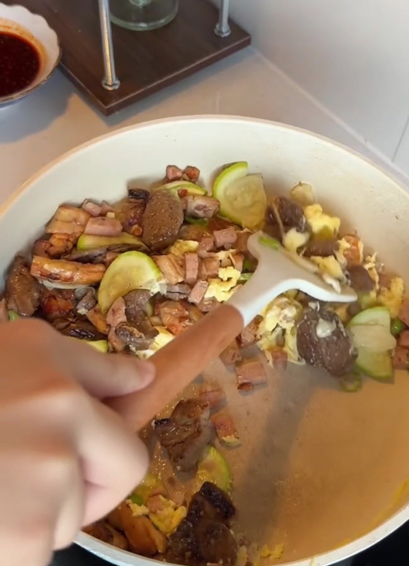 A bowl of white rice being added to a pan containing stir-fried zucchini, meat, and eggs.