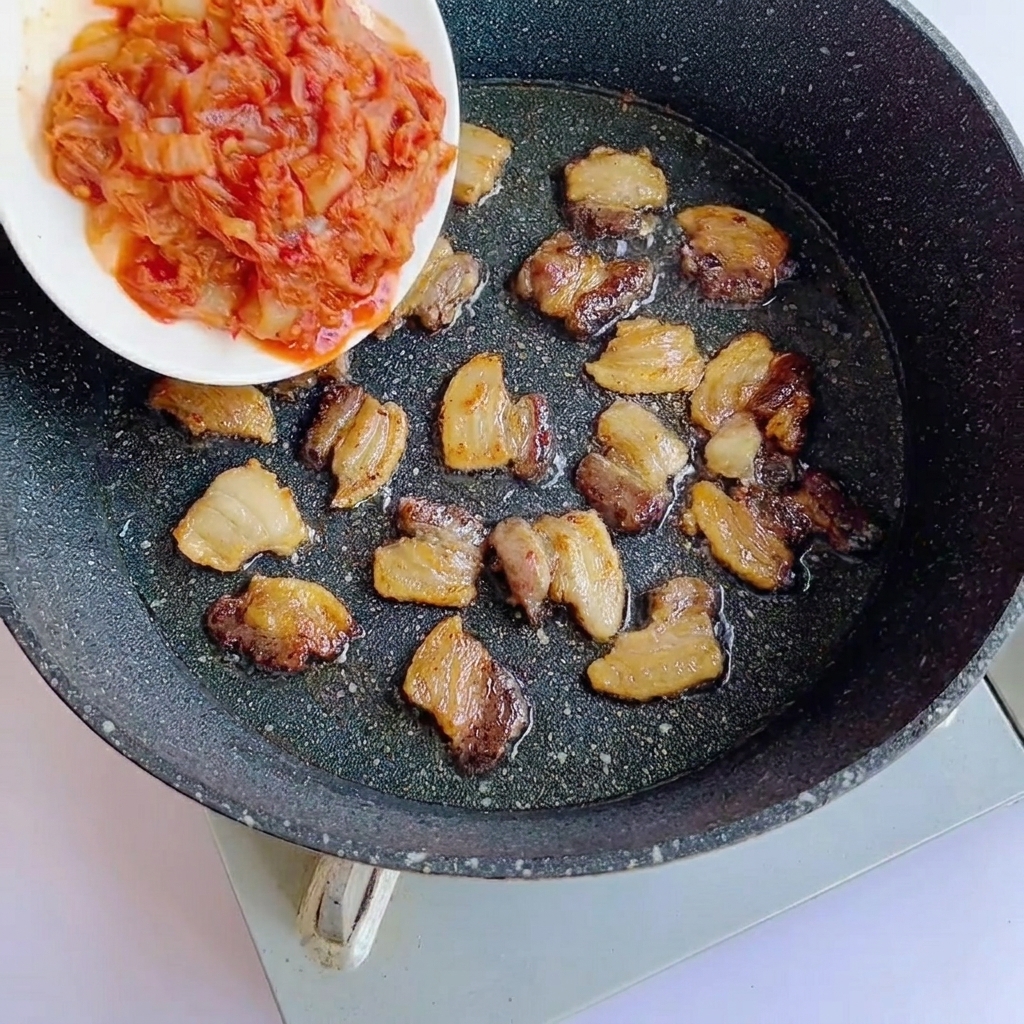 Chopped kimchi being added to a pan containing browned pork belly slices.