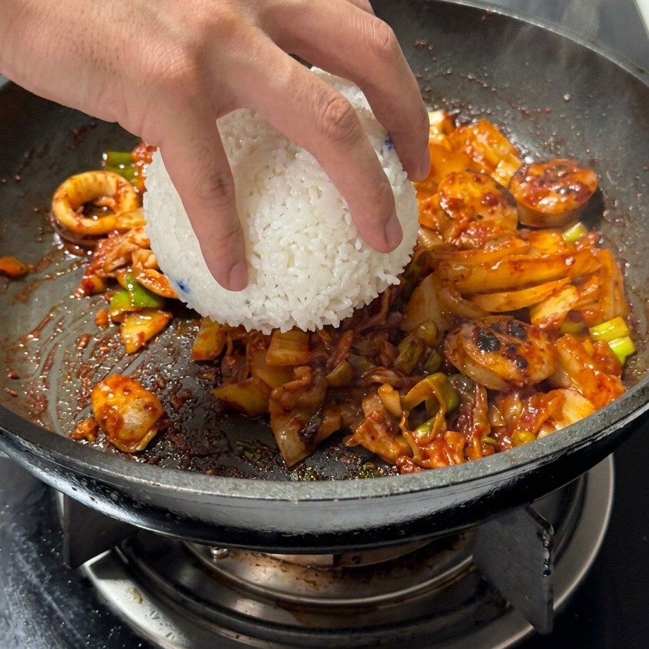 A bowl of white steamed rice being added to a pan containing stir-fried kimchi and meat.