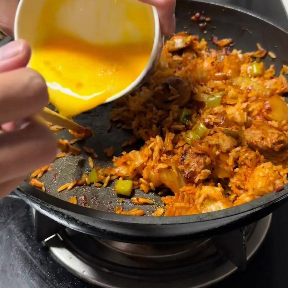Beaten egg being poured into a cleared side of the pan, next to the fried rice mixture.