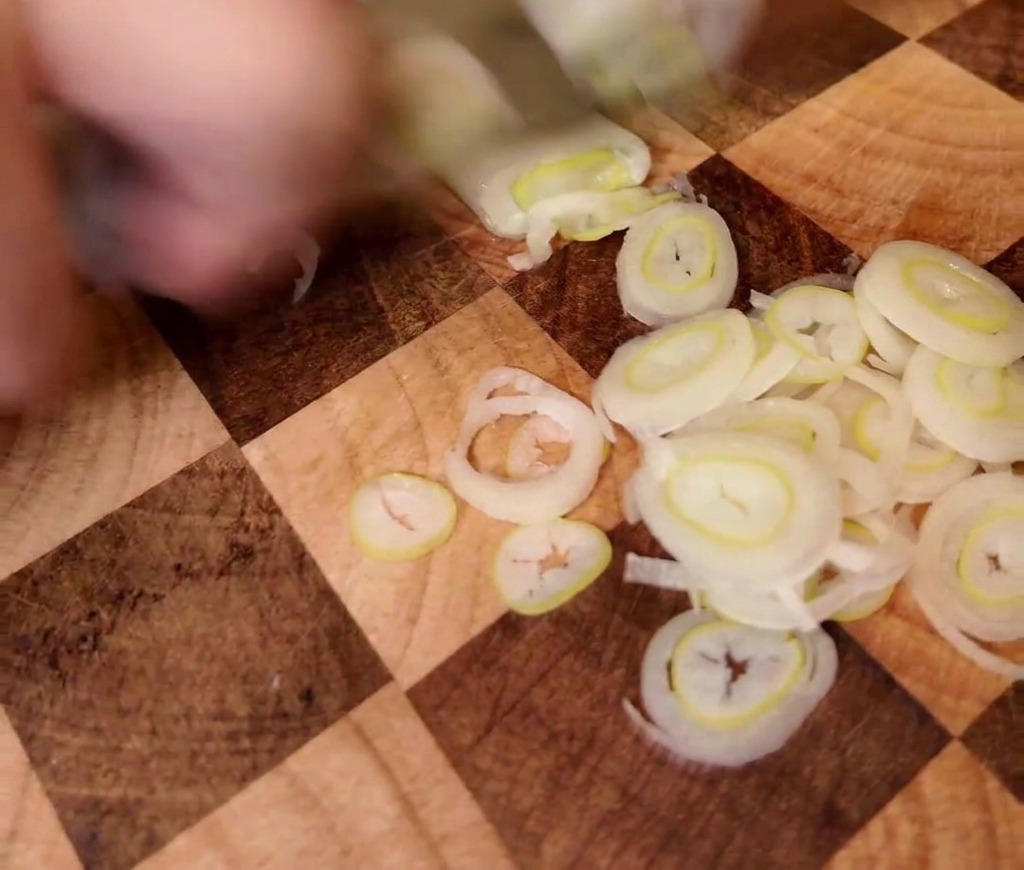 Green onions being sliced thinly on a wooden surface.