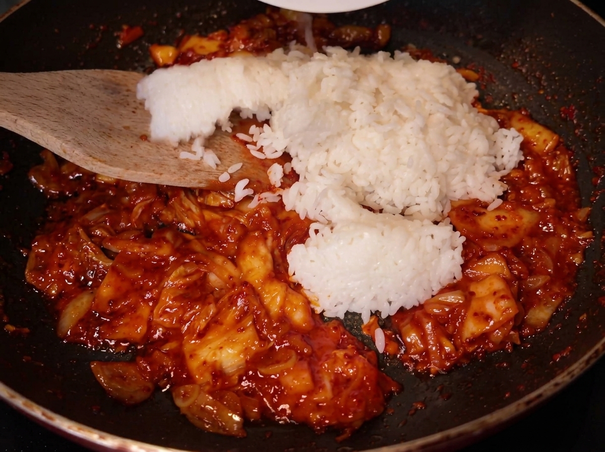 A wooden spatula adding a large scoop of white rice into the pan where the red kimchi mixture is already sautéing.