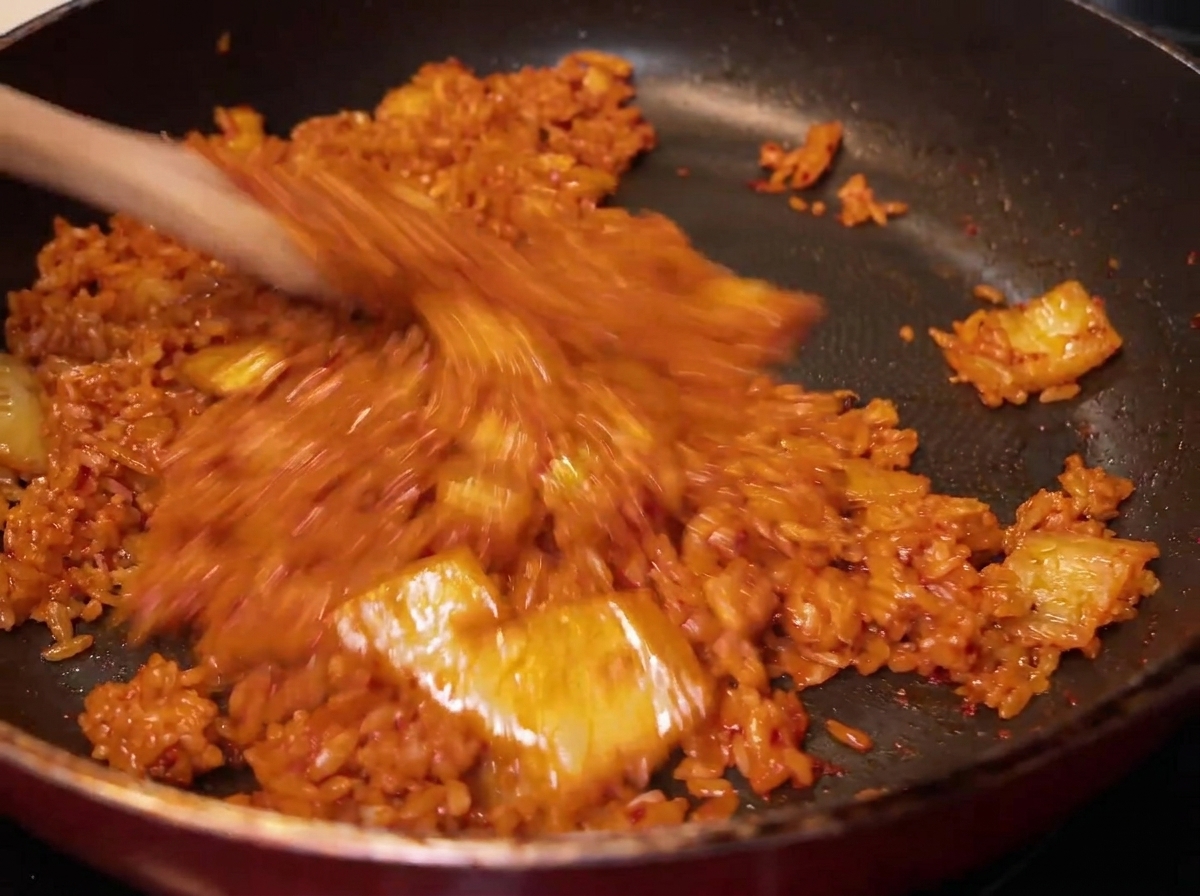 A close-up shot of the rice being tossed and mixed with the red kimchi in the pan, showing the rice turning a uniform reddish-orange color.