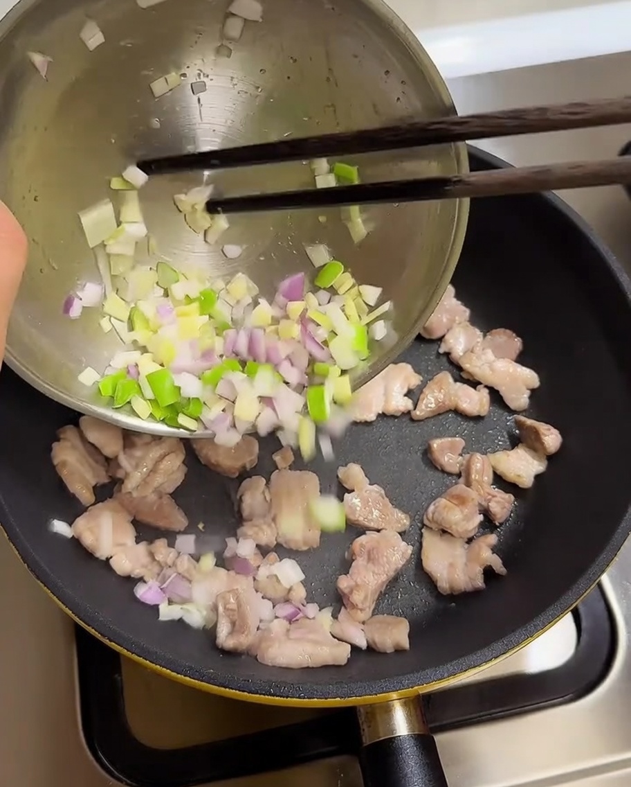 Chopped scallions, garlic, and onions being poured from a bowl into a pan with cooked pork belly.