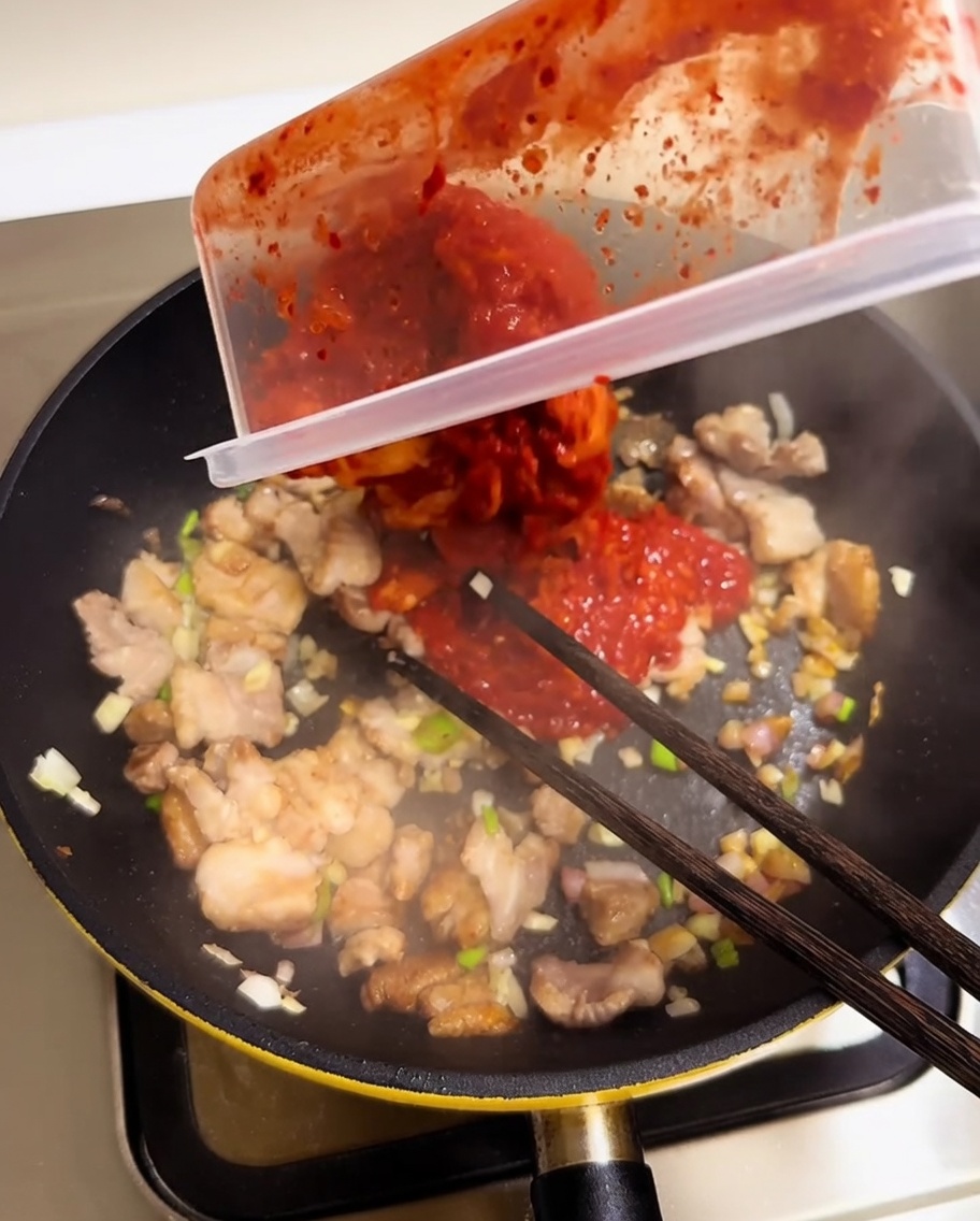Chopped red kimchi being poured from a container into a pan with pork and aromatics.
