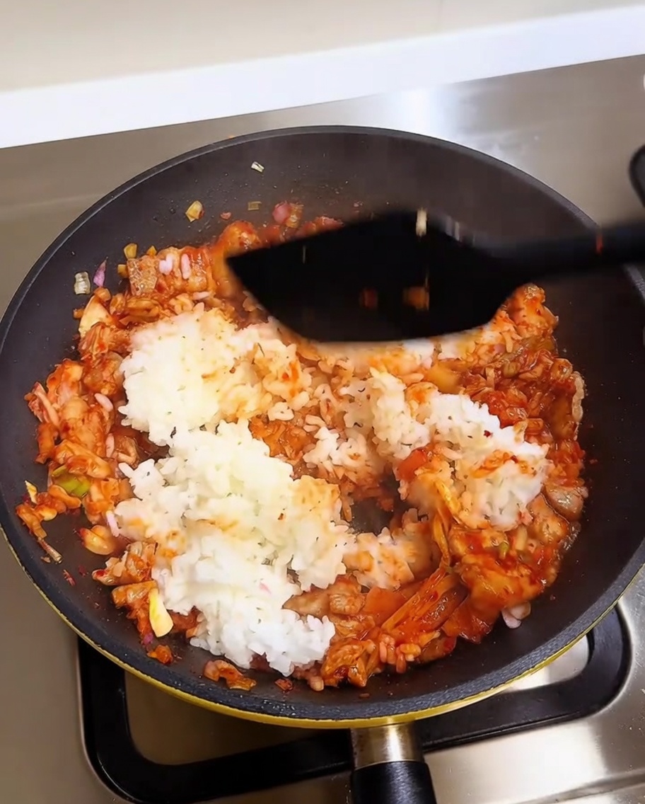 A spatula mixing white rice into a vibrant red pork and kimchi base in a hot pan.