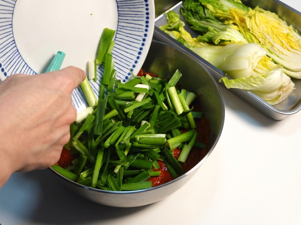 Freshly chopped green chives being added to a thick red kimchi seasoning paste in a metal bowl.