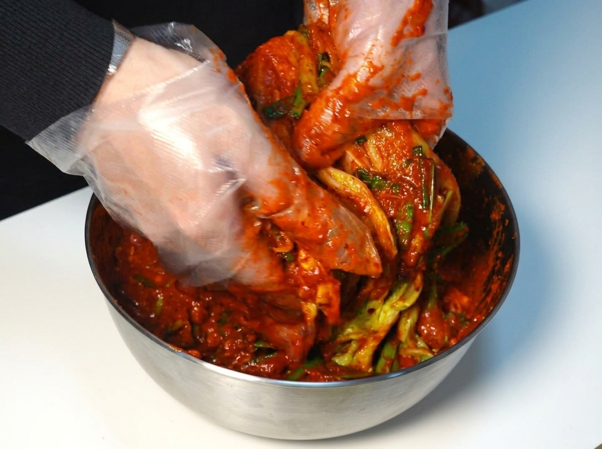 Gloved hands spreading thick red kimchi paste evenly over each layer of a napa cabbage head.