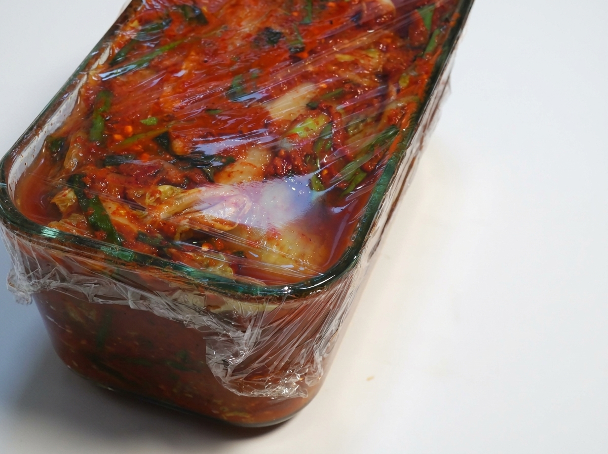 Container of fermented kimchi with the plastic wrap being peeled back.