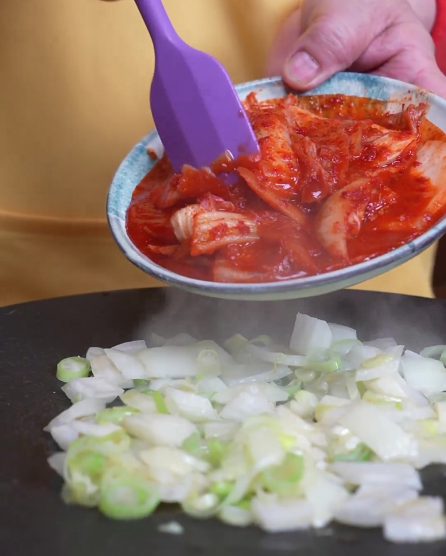 Kimchi being added to the pan where green onions are already cooking.