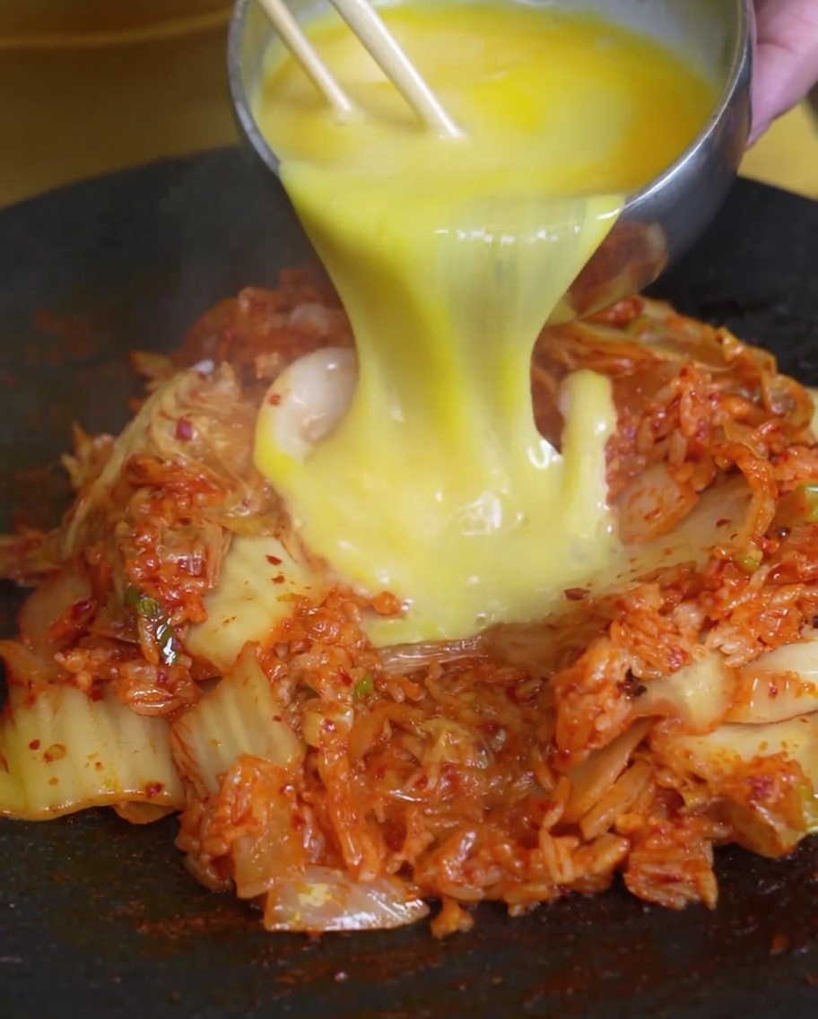 Beaten eggs being poured over a mound of kimchi fried rice in a hot pan.