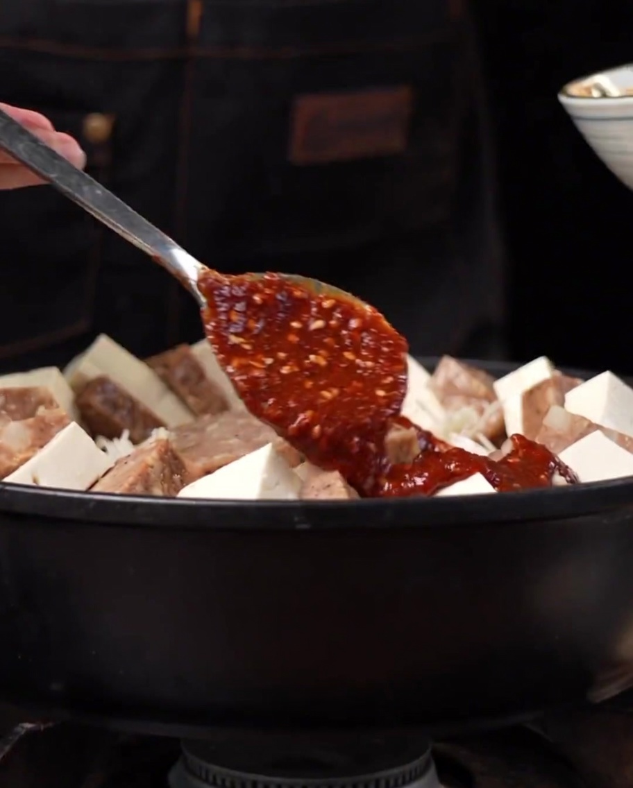 A thick reddish spicy sauce being spooned and spread over arranged ingredients in a metal pot.