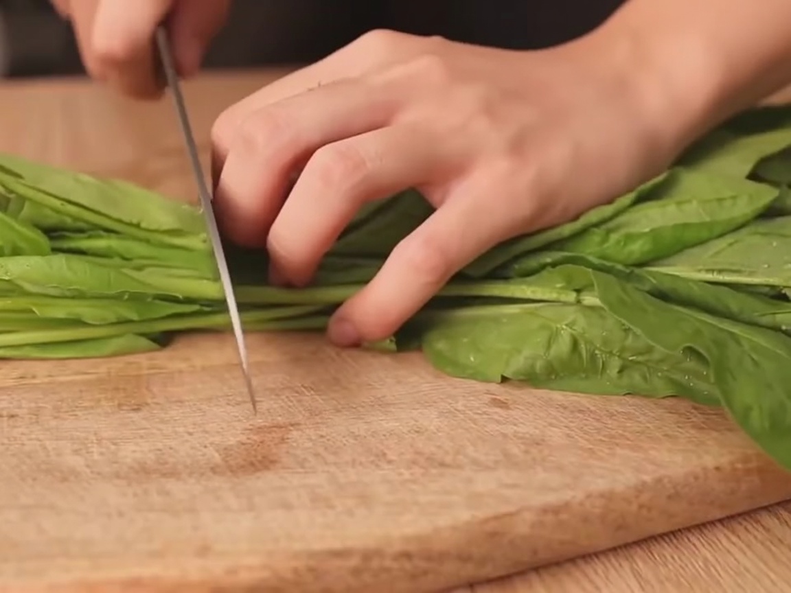A hand cutting a bundle of fresh spinach into sections on a wooden cutting board.