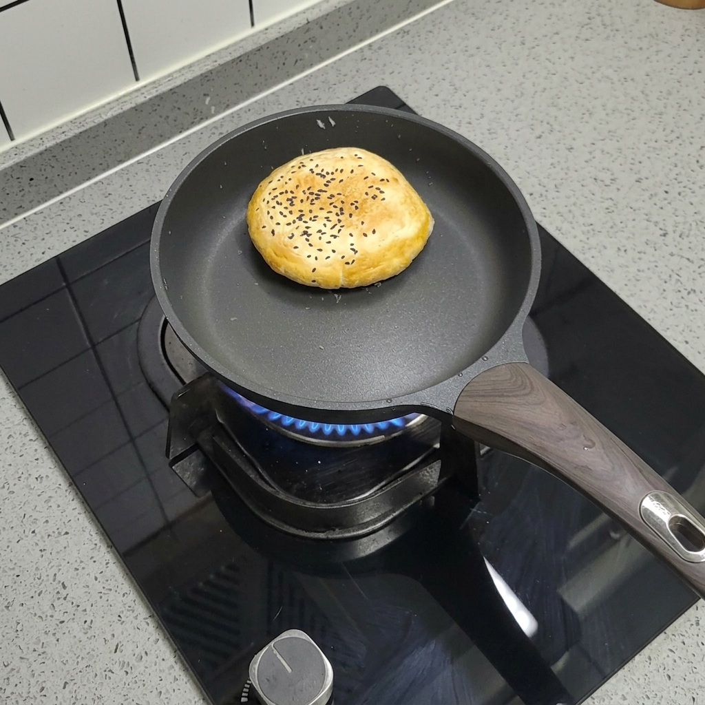 A frozen burger bun with sesame seeds cooking in a dry, non-stick pan on the stove.