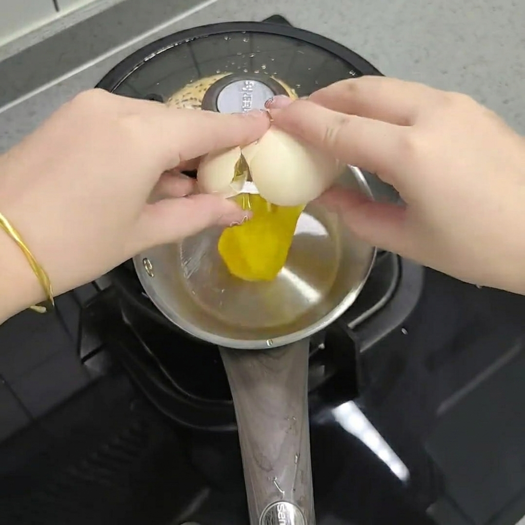 Hands cracking an egg into a small metal pot containing chili oil, powder, sesame salt, and frozen green onions.