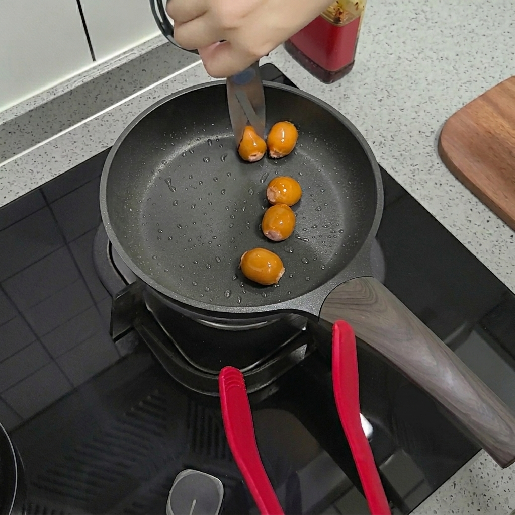 Scissors cutting open small, browned sausages directly in the frying pan.