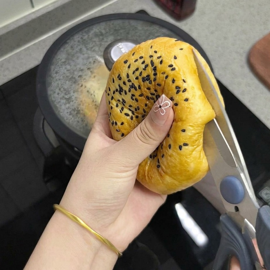A close-up view of a hand using kitchen scissors to cut a toasted, sesame-topped bun in half.