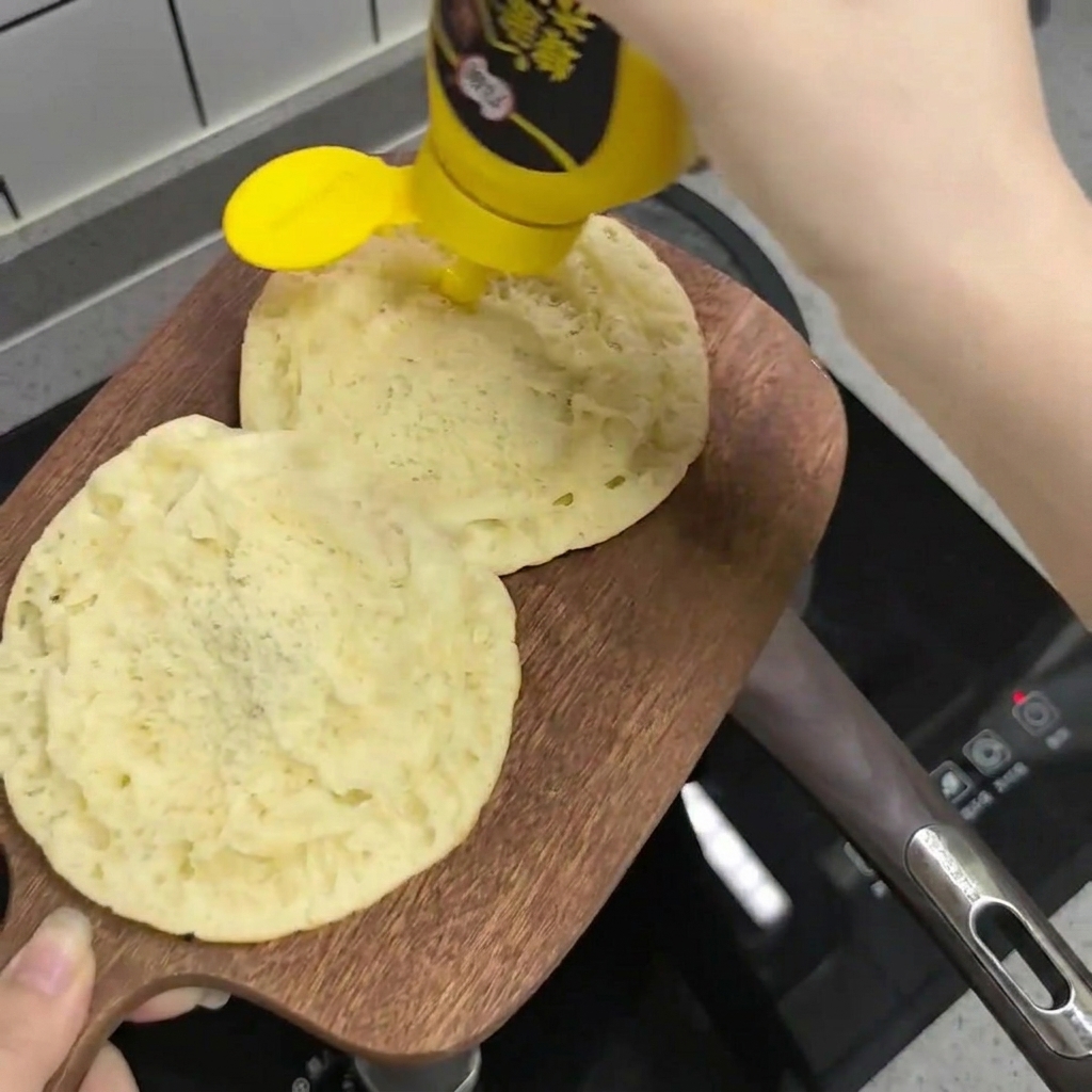 Honey mustard sauce being squeezed from a bottle onto the cut sides of two round burger buns placed on a wooden board.