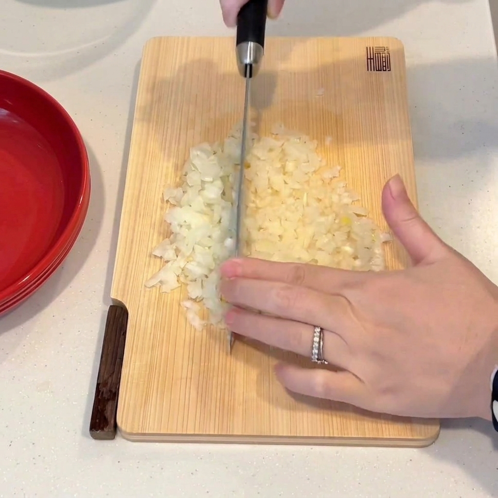 A hand using a chefs knife to finely chop an onion on a wooden cutting board.